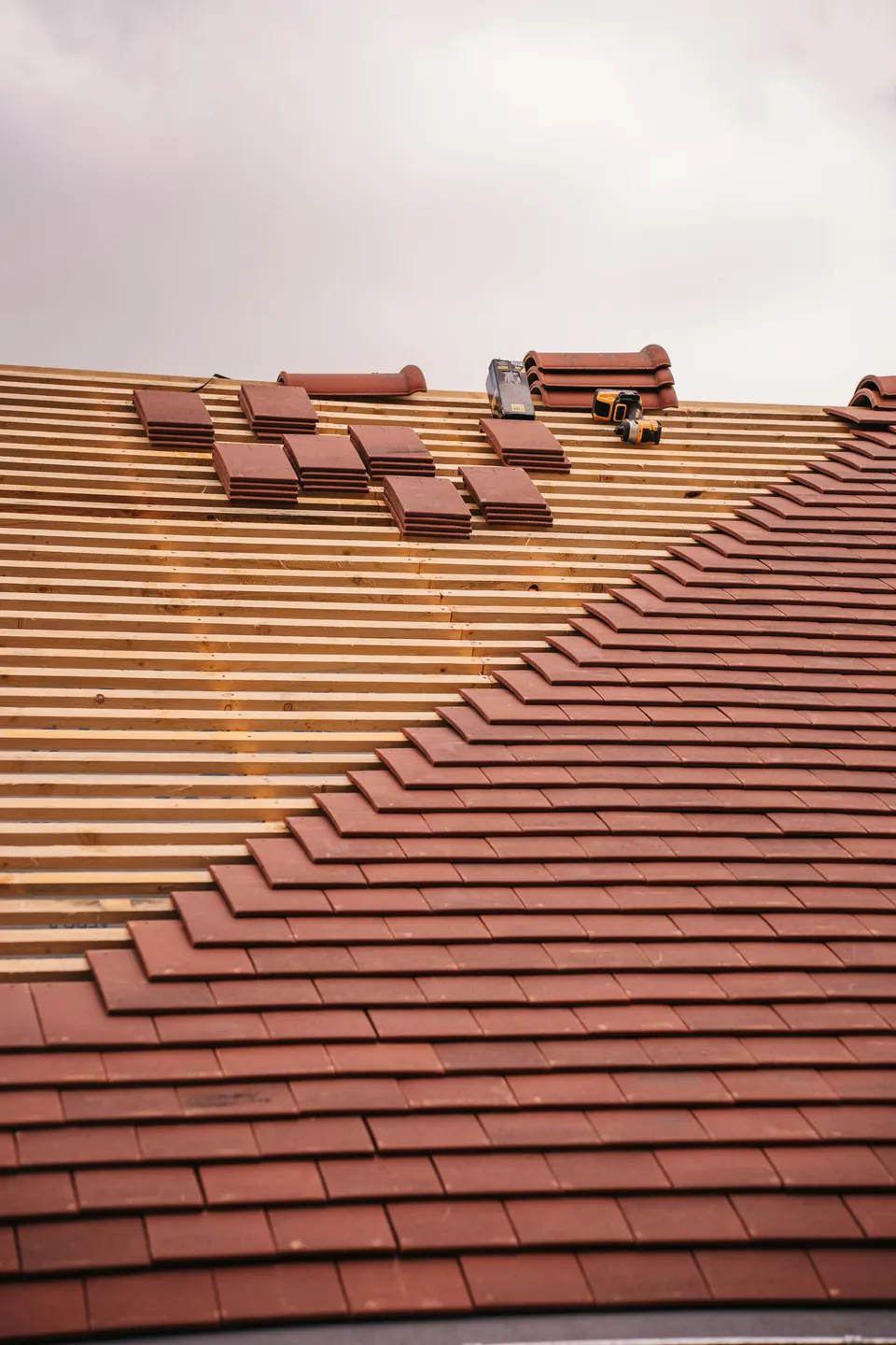 Red plain roof tiles being laid in courses over timber battens, with stacks of tiles further up the roof